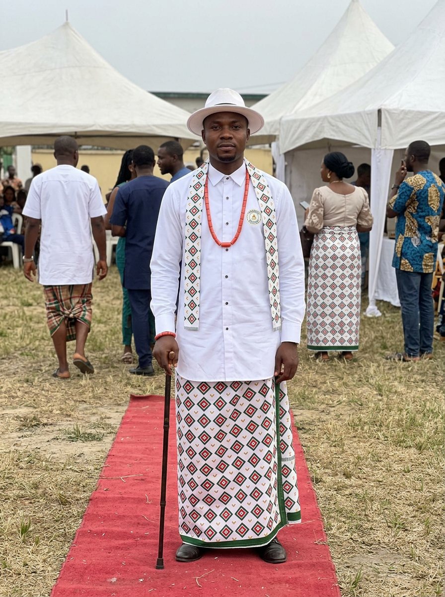 A man on the red carpet in the white festival shirt, diamond-print wrapper and stole, with a hat and walking stick.