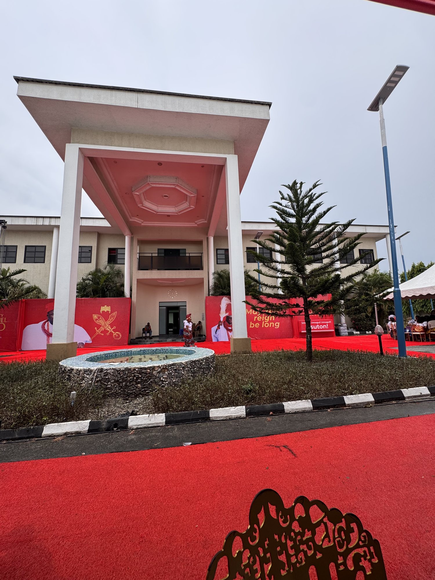 The front of the Palace of Uvwie, a cream building with a tall white portico, a fountain and red carpet, with red royal banners along the forecourt.