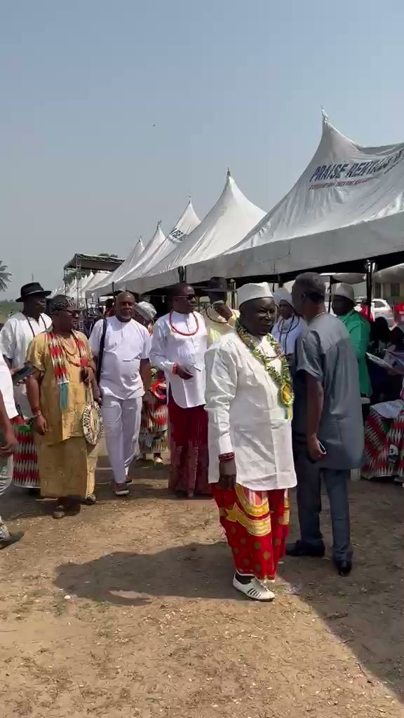 Chiefs in white shirts over tied wrappers with coral bead necklaces walking past the tents.